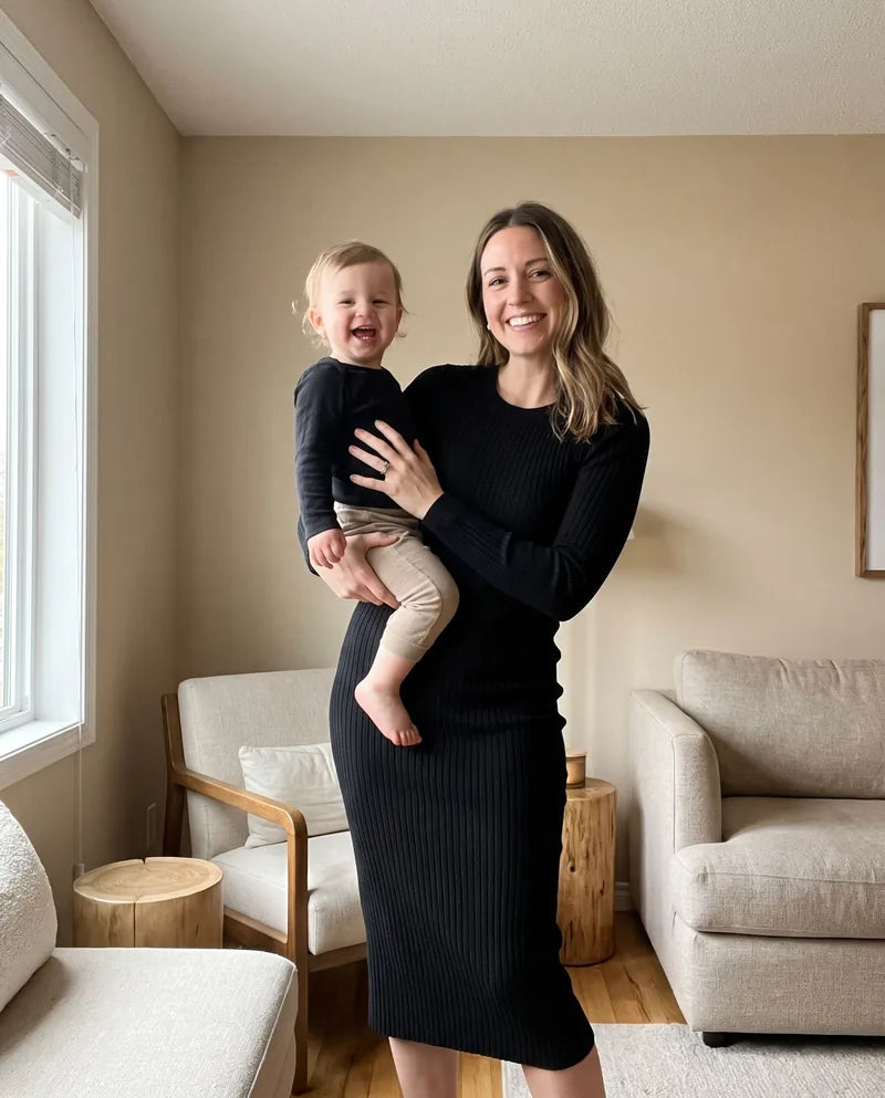 A smiling mom in a fitted black dress, flat belly, holding her toddler on her hip